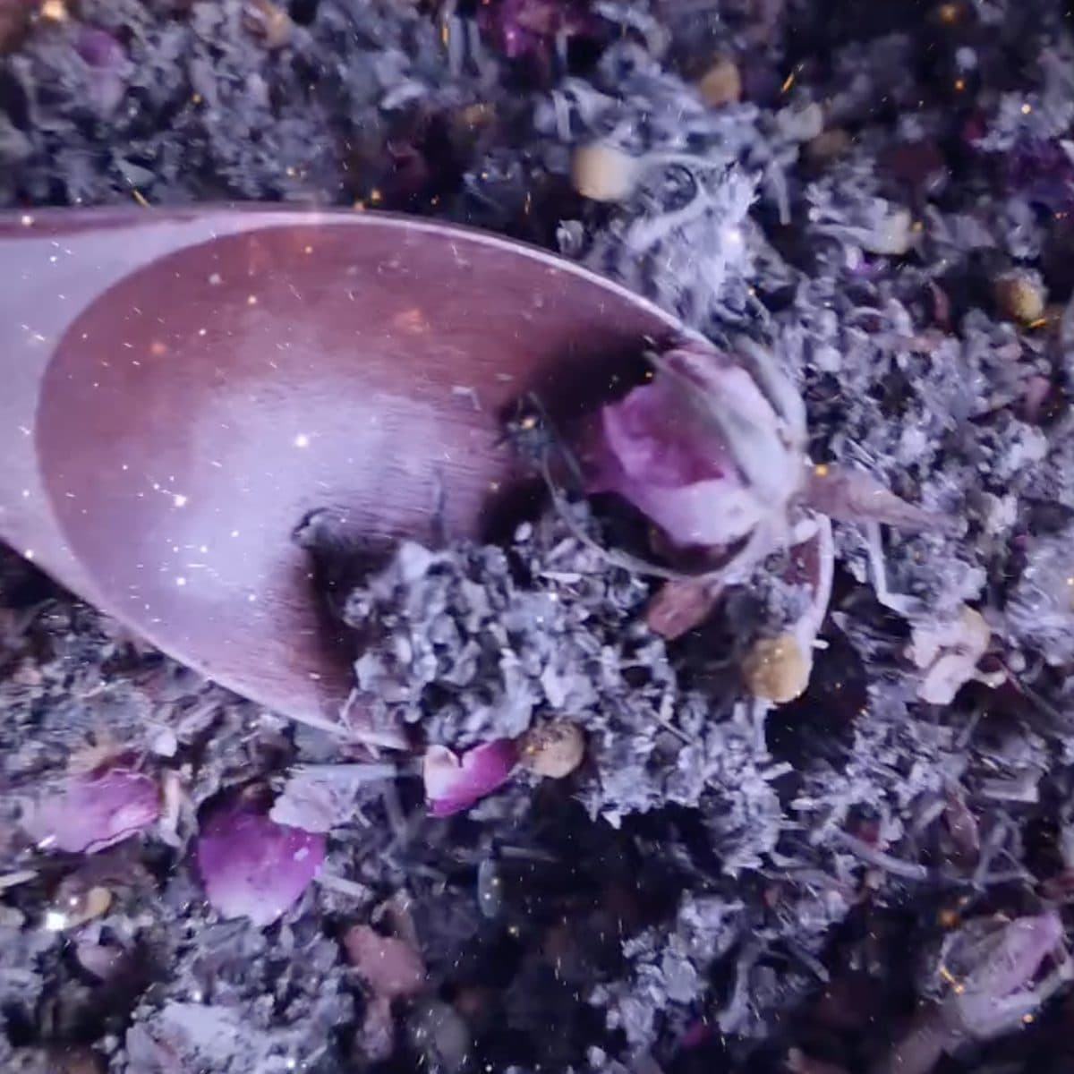 Wooden spoon stirring a glittering herbal tea blend with rose petals and amber resin